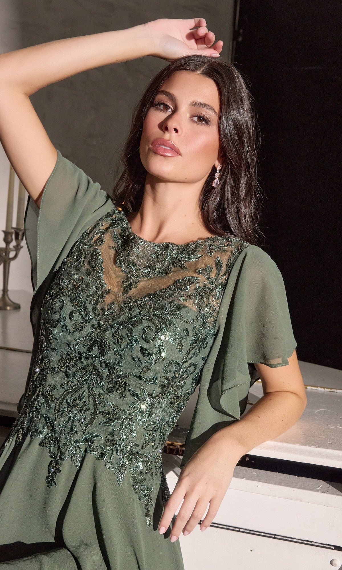 A woman in the Ladivine CL09 Long Formal Dress, adorned with green sequins, poses indoors with one arm on a white surface and the other raised over her head.